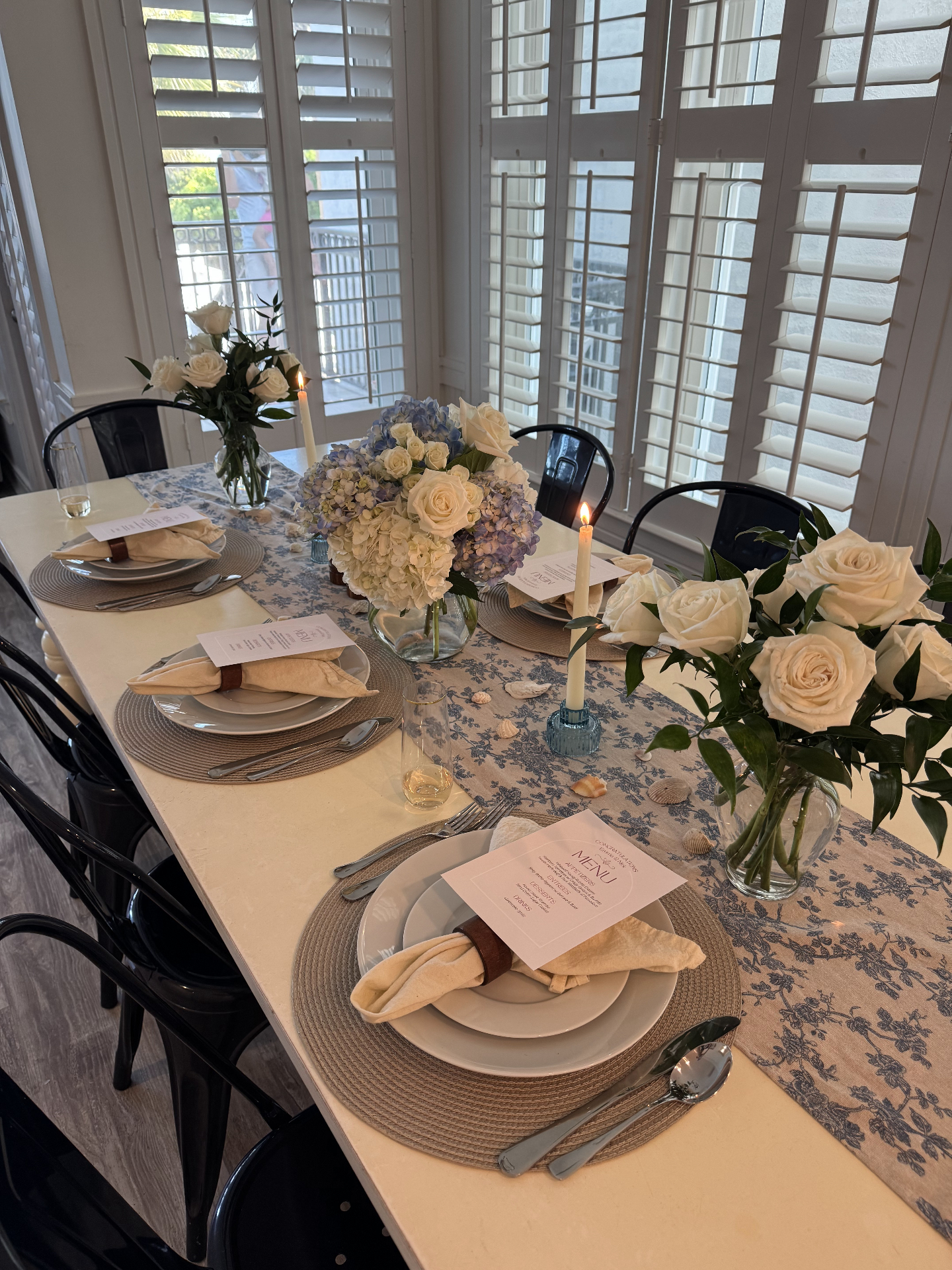 An elegant vacation rental table set with blue toile linens, white roses, hydrangeas, and taper candles